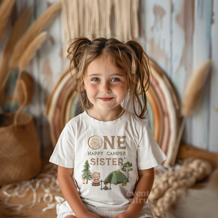 Smiling girl with pigtails in front of a boho rainbow in a white One Happy Camper Sister shirt.