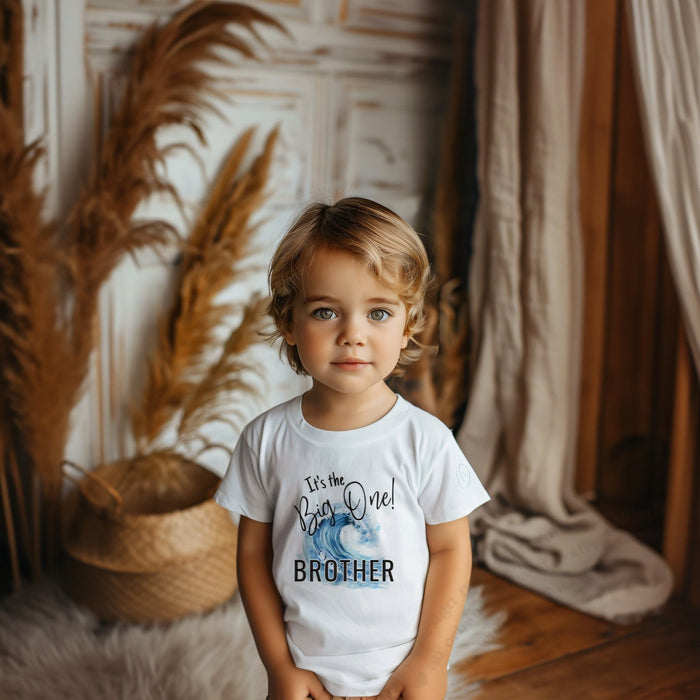 Little boy in a white It's the Big One Brother shirt with a blue wave.