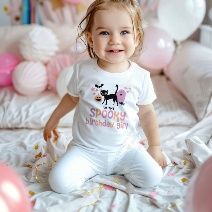 Smiling toddler girl among pink balloons in a white I'm the Spooky Birthday Girl tee.