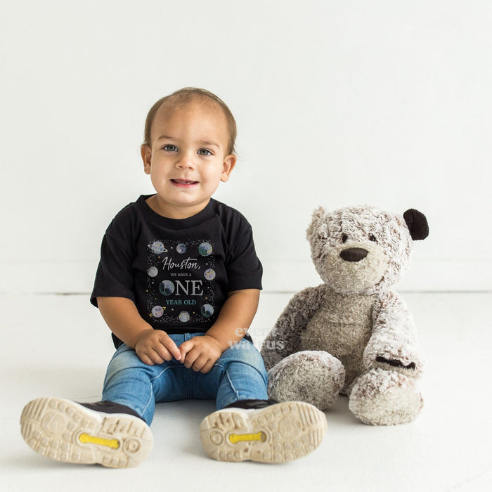 Toddler sitting by a teddy bear in a black Houston We Have a One Year Old shirt.