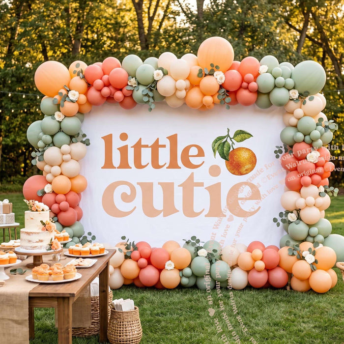 Little cutie backdrop in a garden under an orange, peach and sage balloon garland beside a dessert table.
