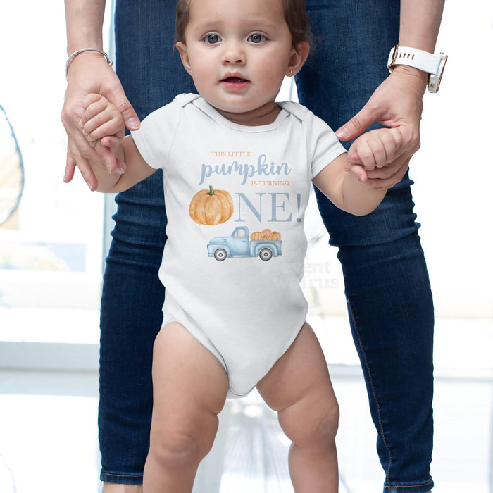 Baby taking steps while holding Mom's hands, in a white This Little Pumpkin Is Turning One bodysuit.