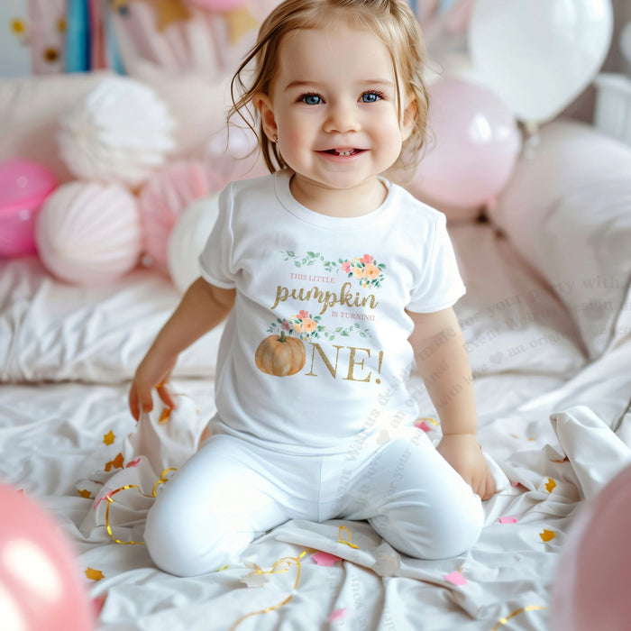 Smiling toddler girl among pink balloons in a white This Little Pumpkin Is Turning One tee.