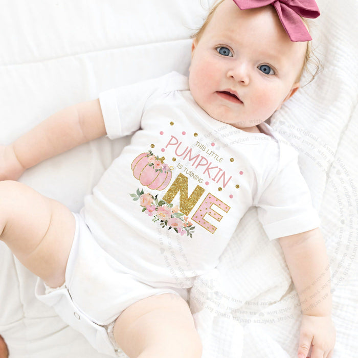 Baby girl with a pink bow lying in a white This Little Pumpkin Is Turning One bodysuit.