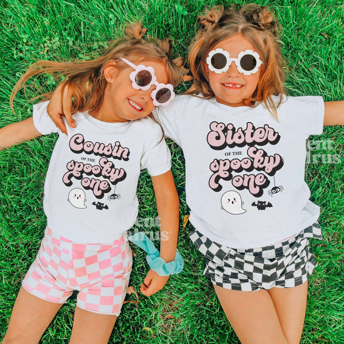 Two girls in heart sunglasses lying on grass in white Cousin and Sister Spooky One shirts.