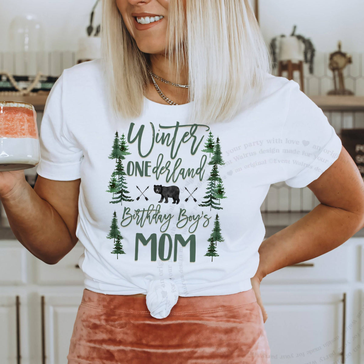 Woman in a blush skirt wearing a white Winter Onederland Birthday Boy's Mom shirt with green pines and a bear.