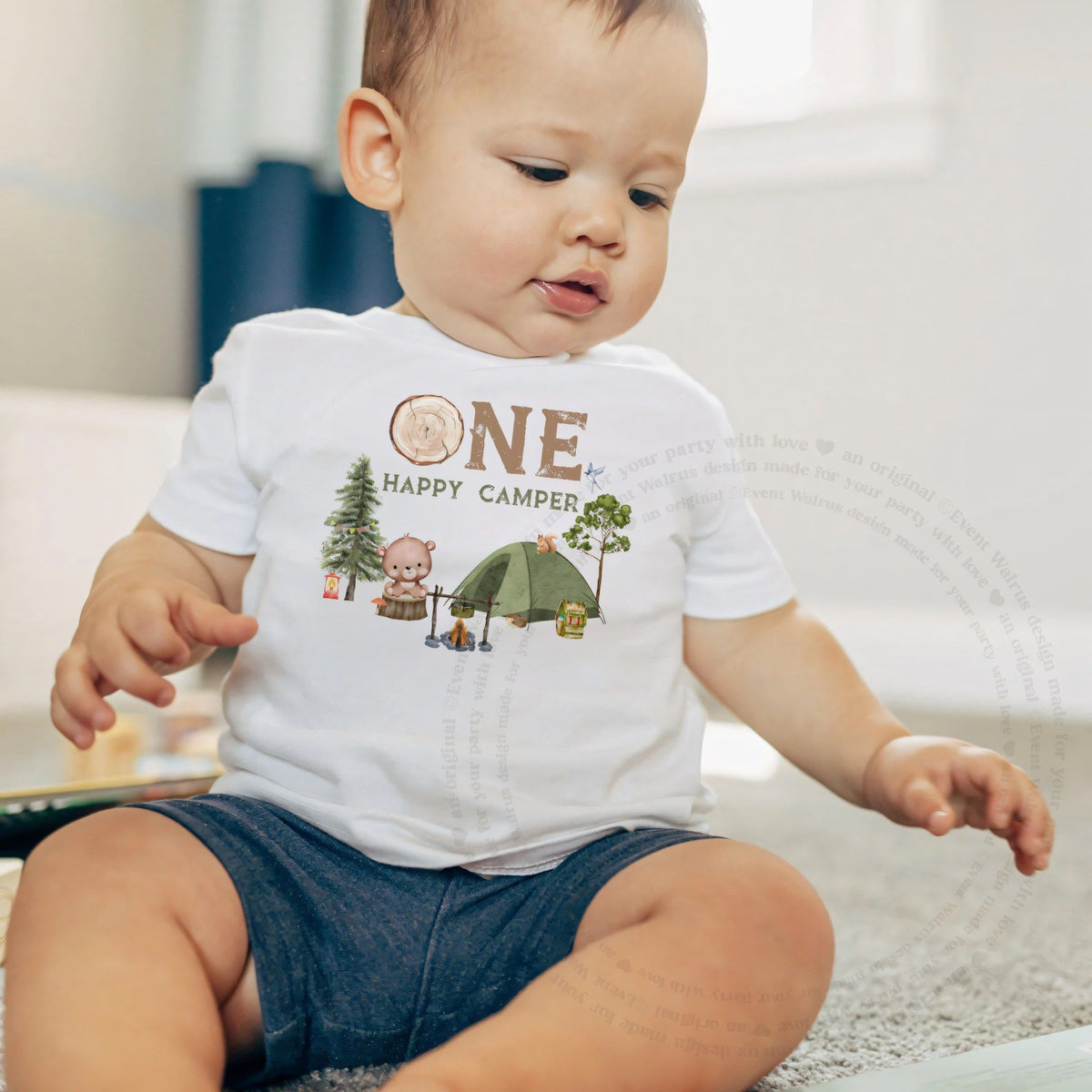 Baby boy sitting on a rug in a white One Happy Camper baby tee.