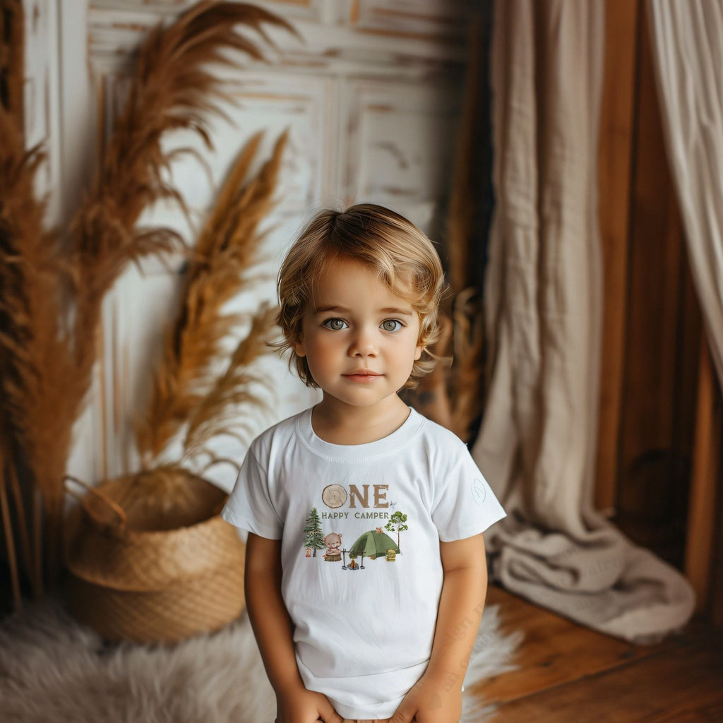 Toddler boy in a white One Happy Camper tee with a bear by a campfire.