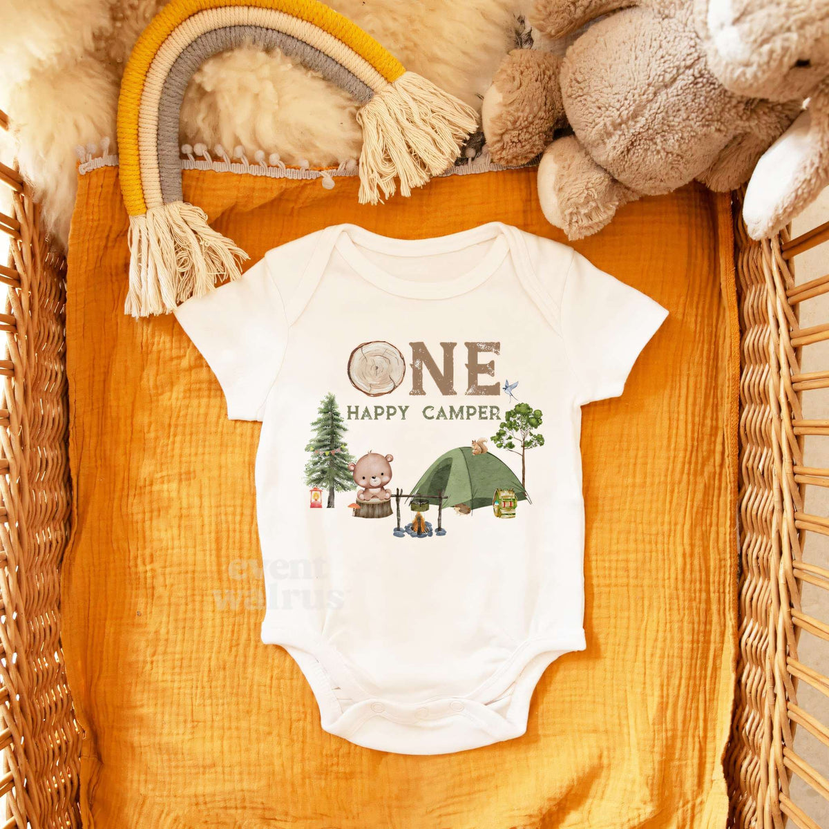 White One Happy Camper bodysuit in a wicker basket with a rainbow toy and a teddy bear.