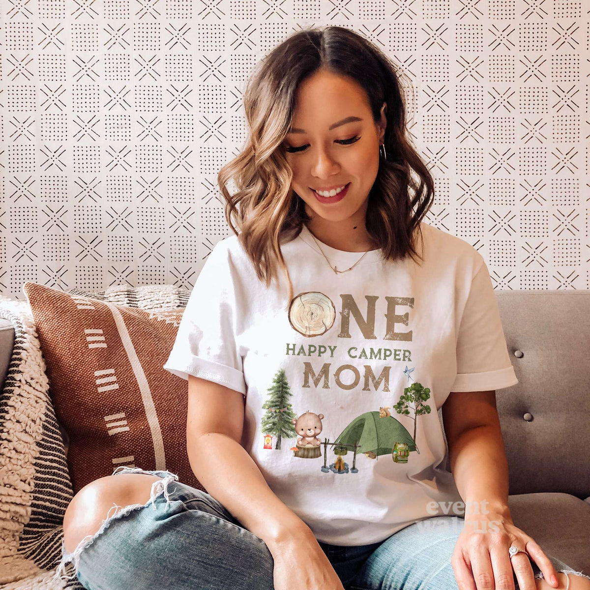 Smiling mom on a sofa in a white One Happy Camper Mom shirt with a bear, campfire and green tent.