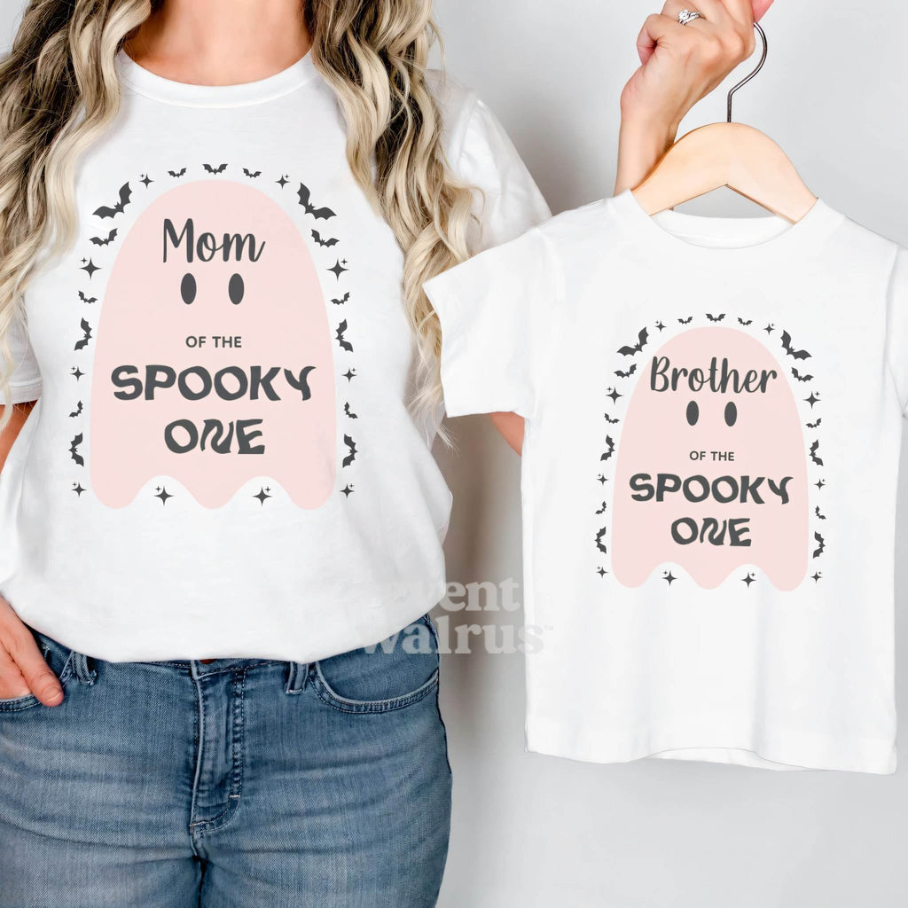 Mom in a white Mom of the Spooky One shirt holding up a matching Brother tee.