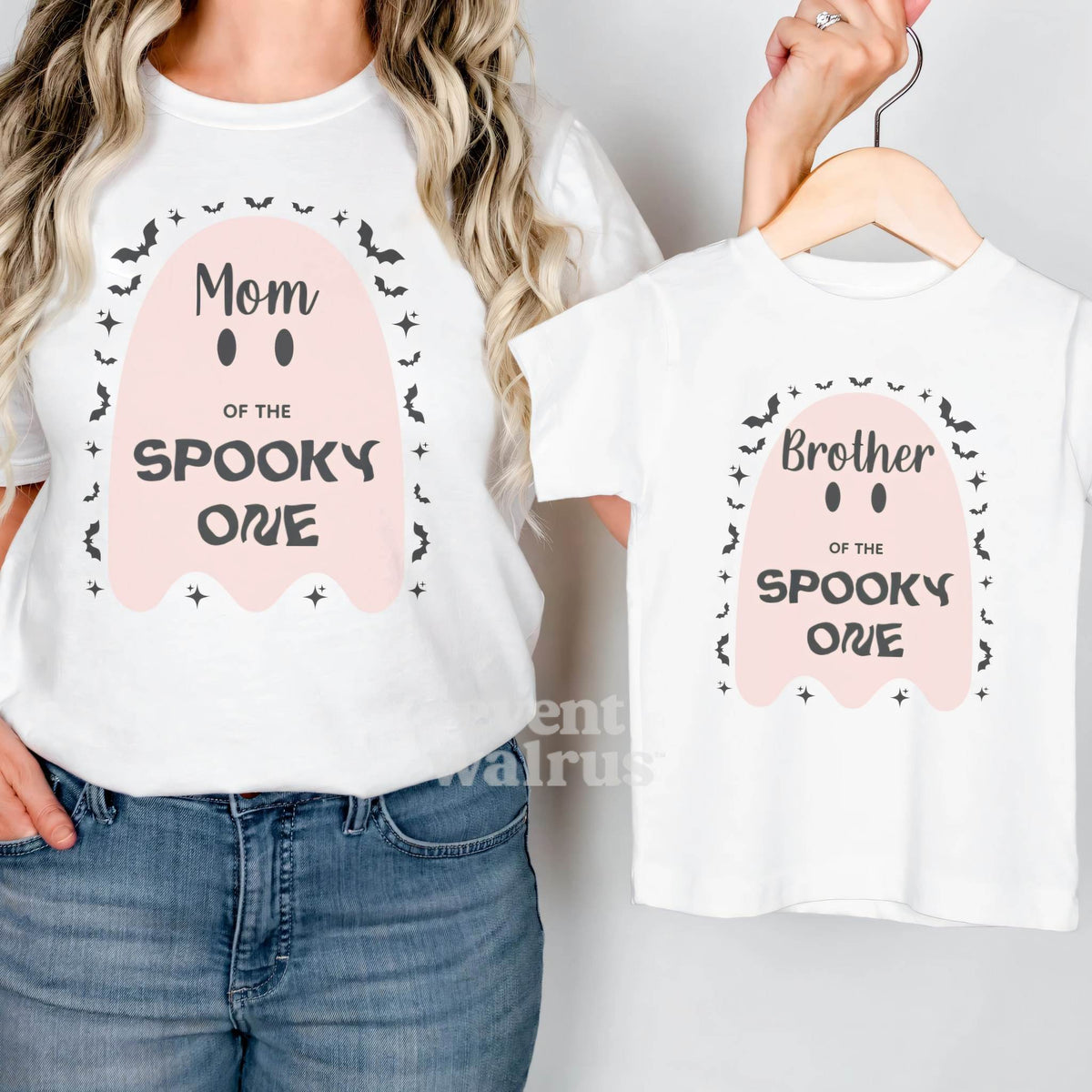 Mom in a white Mom of the Spooky One shirt holding up a matching Brother tee.