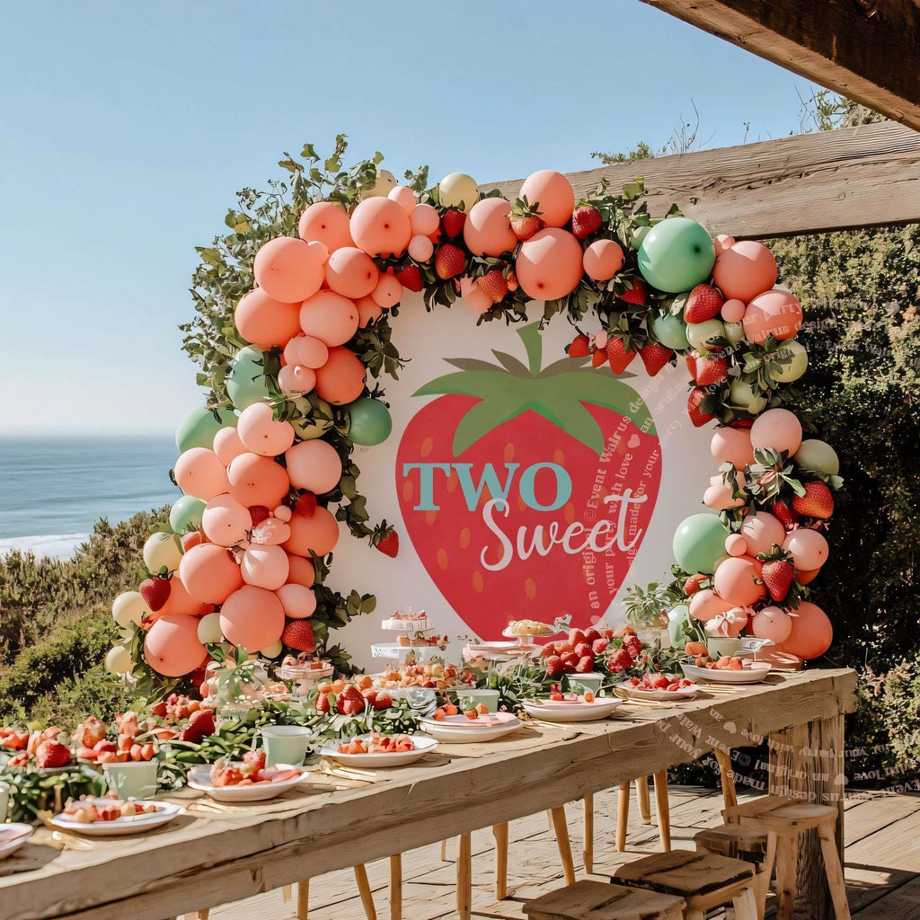 Big strawberry two sweet backdrop above a long seaside table with peach balloons.