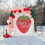 Two sweet big strawberry backdrop in the snow under string lights with a pink and green balloon arch.