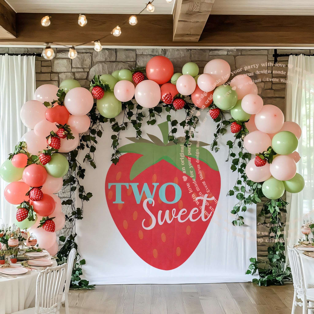 Big strawberry backdrop in a stone room with pink and green balloons and hanging vines.