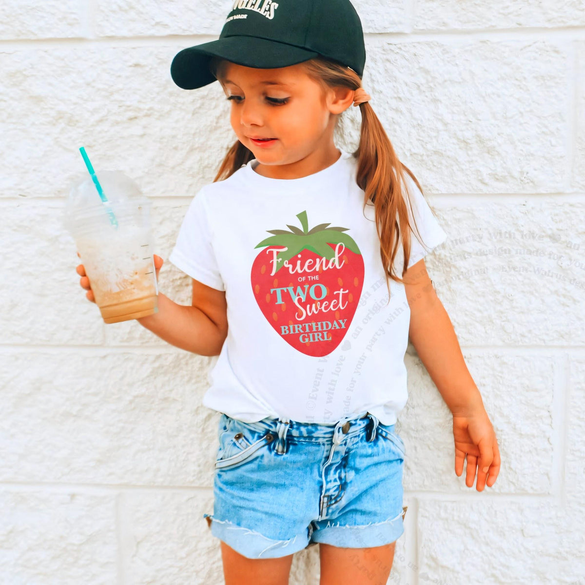 Girl in a ball cap holding a drink in a white Friend of the Two Sweet shirt.