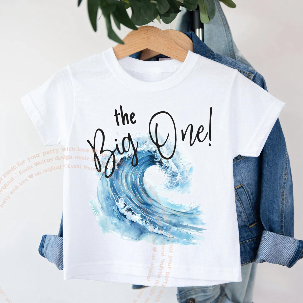 White the Big One toddler shirt with a watercolor wave on a wooden hanger.