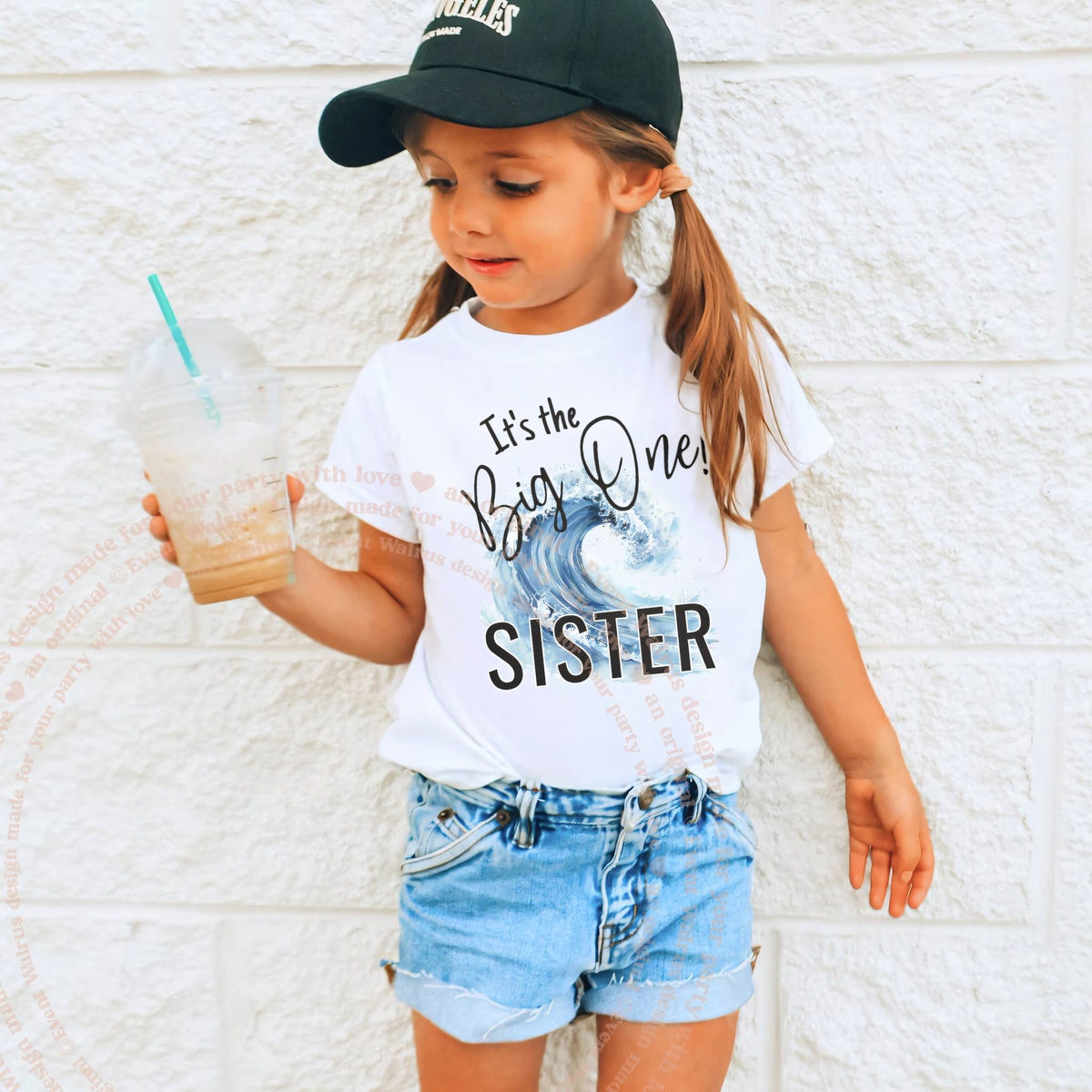 Girl in a ball cap wearing a white It's the Big One Sister wave tee.