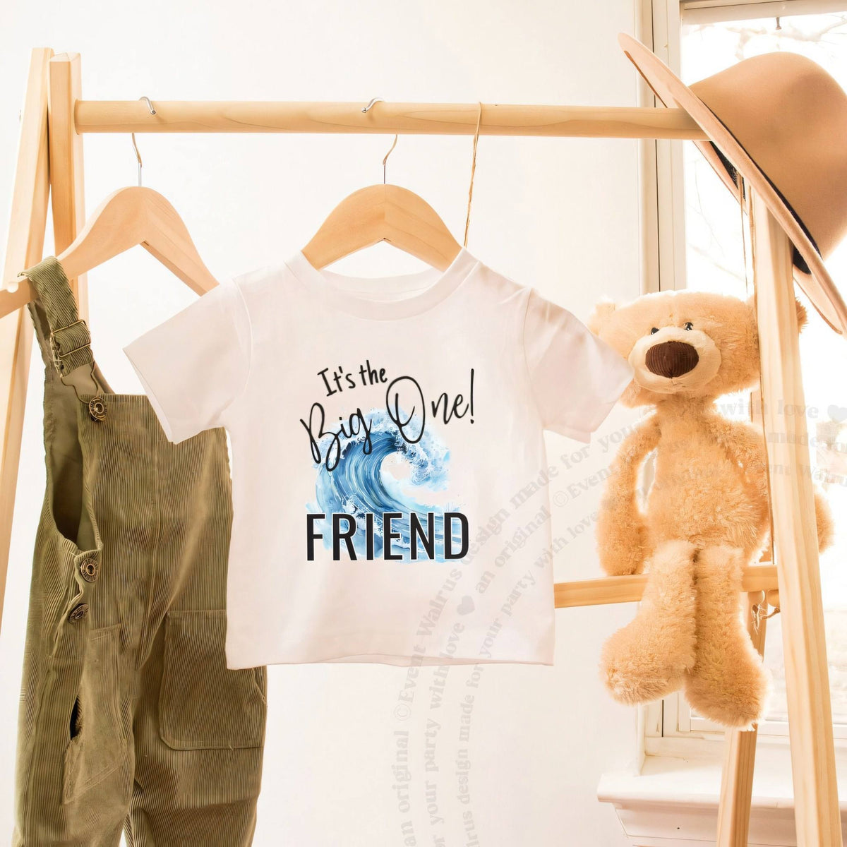 White toddler It's the Big One Friend shirt on a hanger beside a teddy bear.