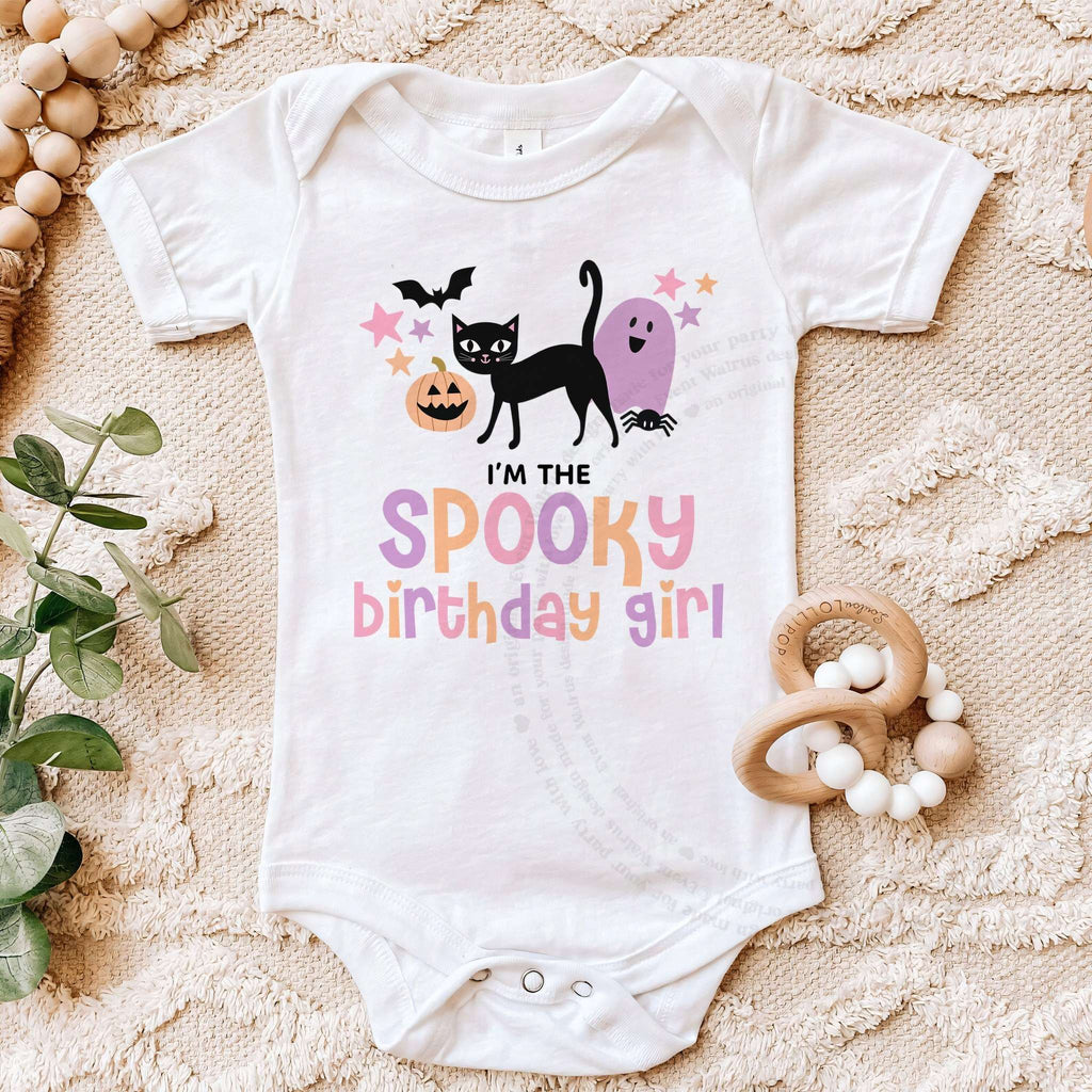 White I'm the Spooky Birthday Girl bodysuit on a beige blanket with a wooden teether.