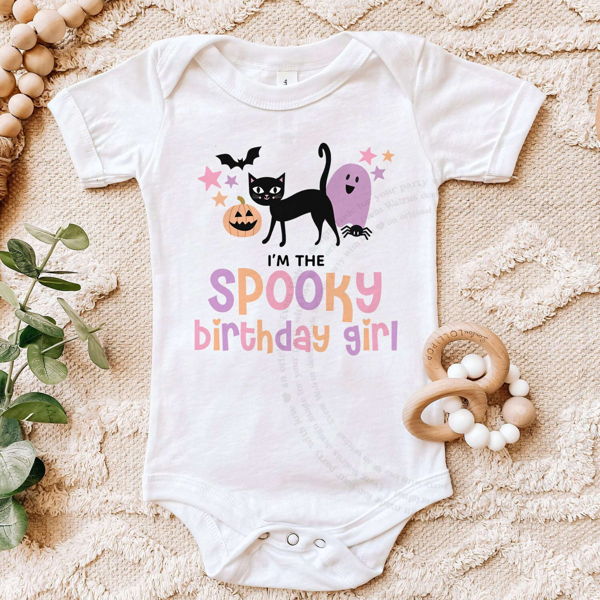 White I'm the Spooky Birthday Girl bodysuit on a beige blanket with a wooden teether.