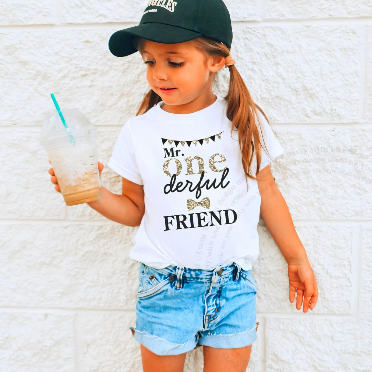Girl in a ball cap holding a drink in a white Mr Onederful Friend shirt.