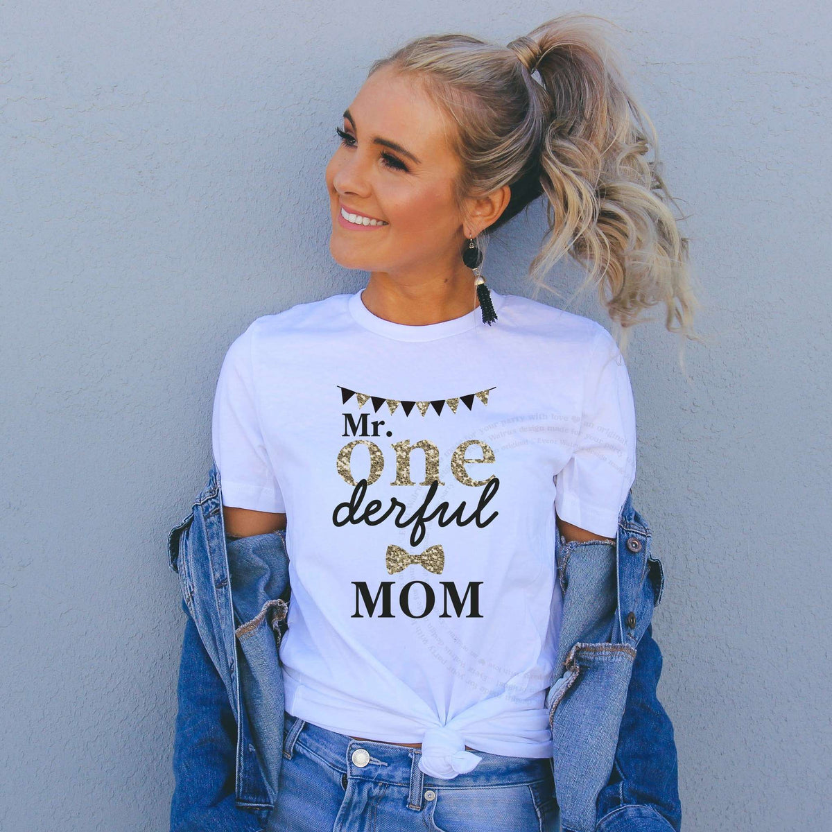 Smiling woman in a denim jacket wearing a white black and gold Mr Onederful Mom shirt.