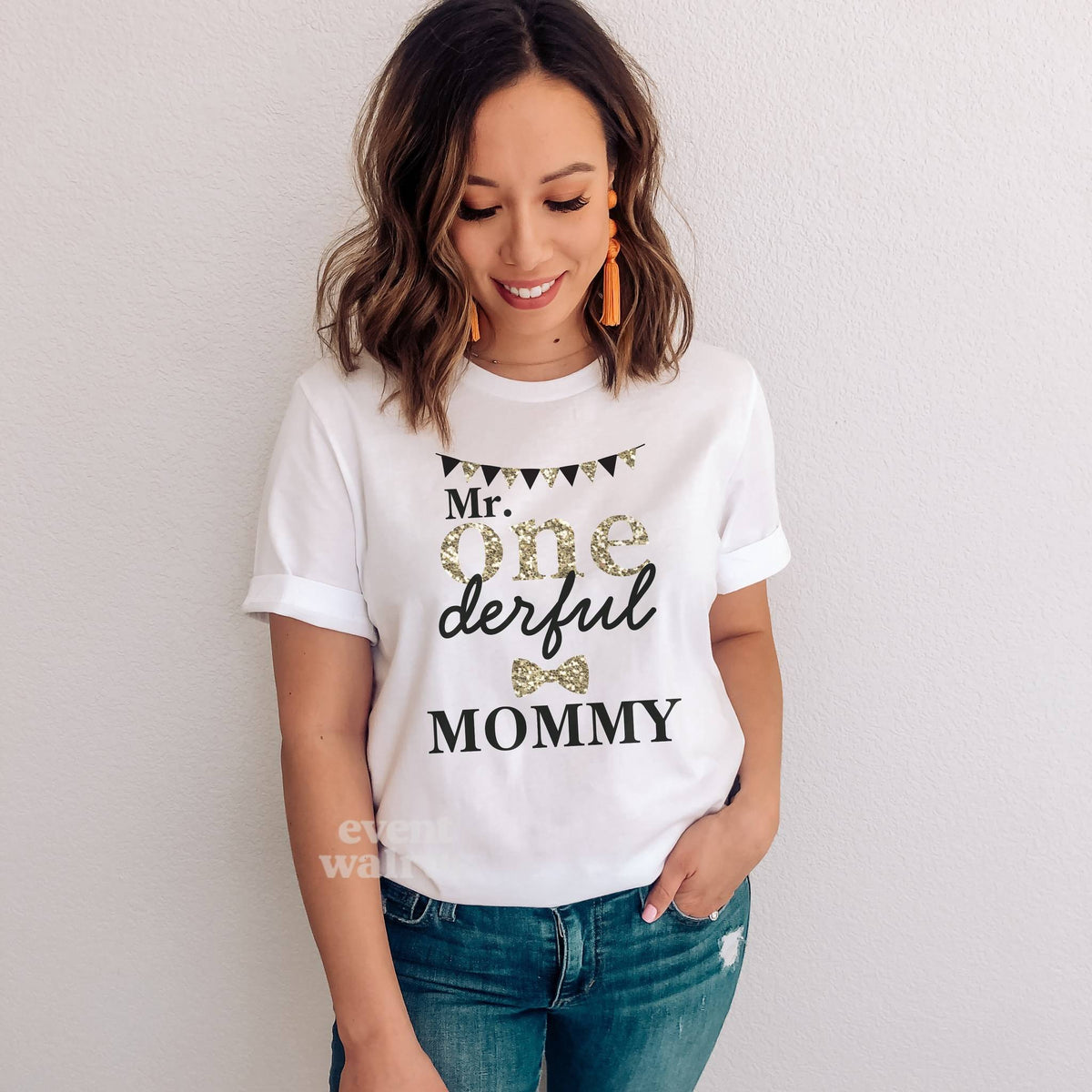 Smiling woman in jeans wearing a white Mr Onederful Mommy shirt with gold glitter ONE and a gold bow tie.