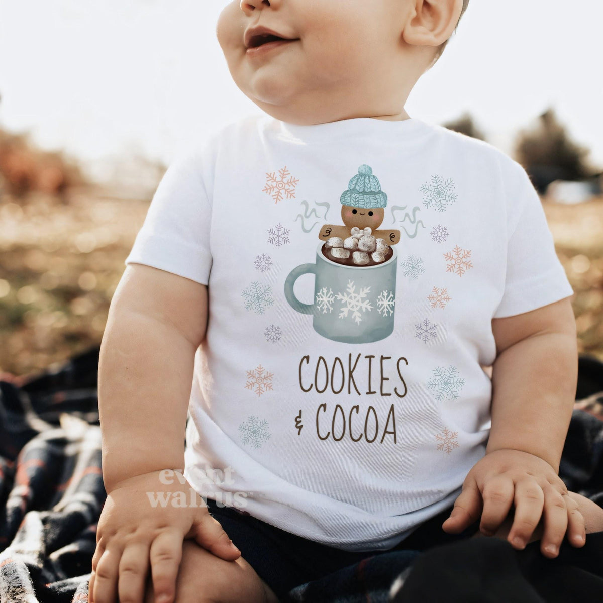 Baby sitting on a blanket outdoors in a white Cookies and Cocoa baby tee.