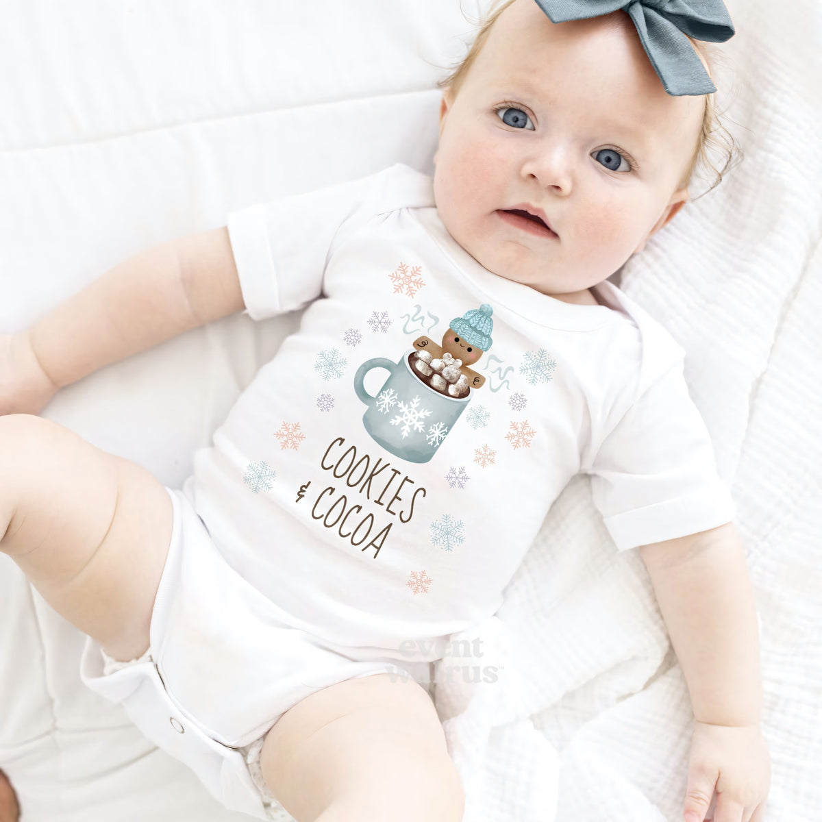 Baby girl with a blue bow lying in a white Cookies and Cocoa bodysuit.