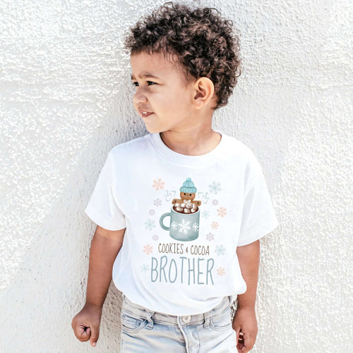 Curly-haired boy against a white wall in a Cookies and Cocoa Brother shirt.