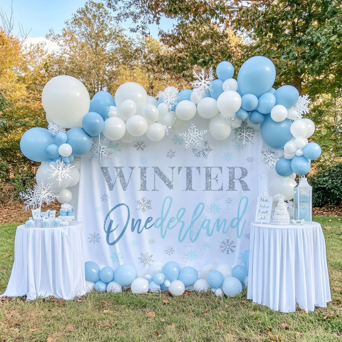 Winter onederland boy backdrop on a lawn with a blue balloon garland and two white dessert tables.
