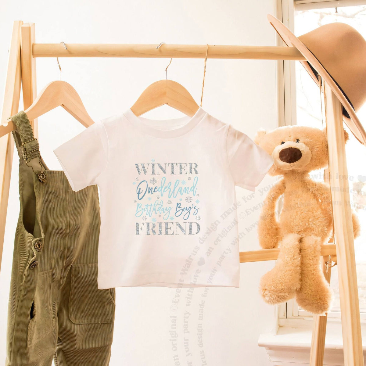 White blue Winter Onederland Friend toddler tee on a wooden clothes rack beside a teddy bear.