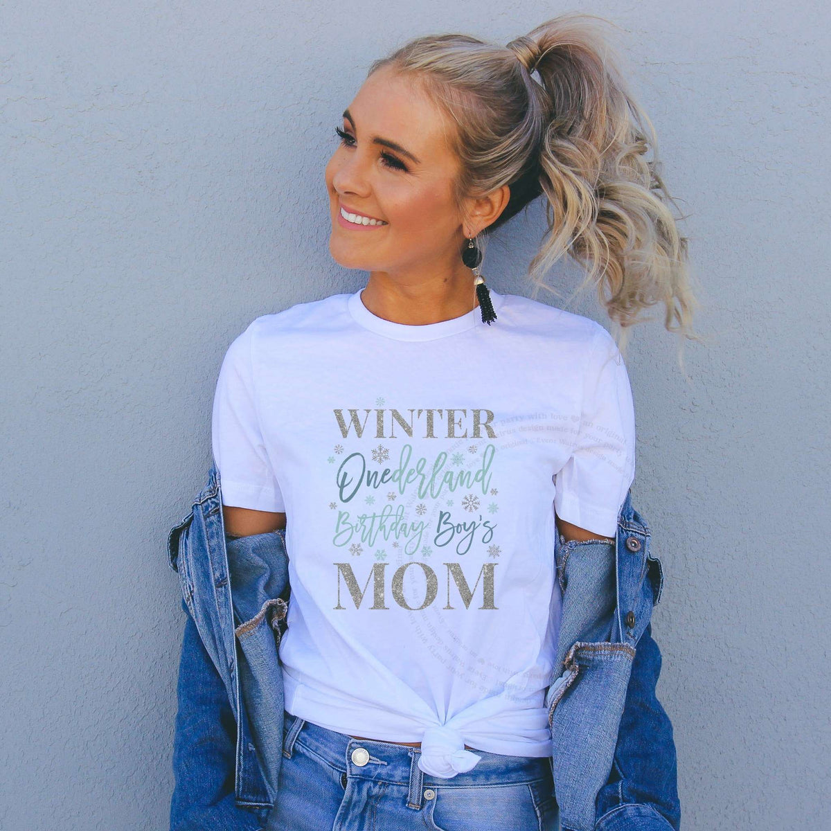 Smiling woman in a denim jacket wearing a white Winter Onederland Birthday Boy's Mom shirt in icy blue and silver.