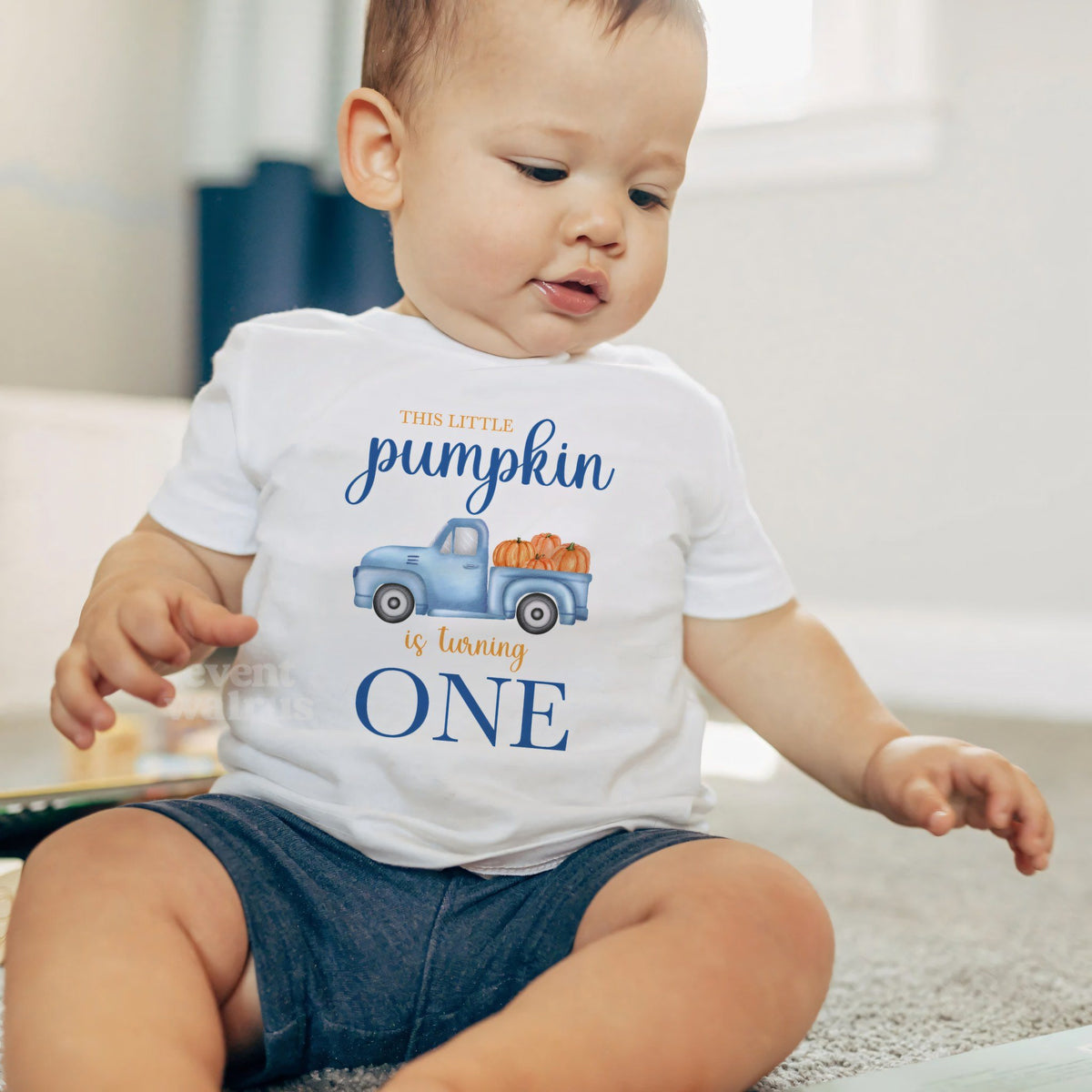 Baby boy sitting on a rug in a white This Little Pumpkin Is Turning One baby tee.