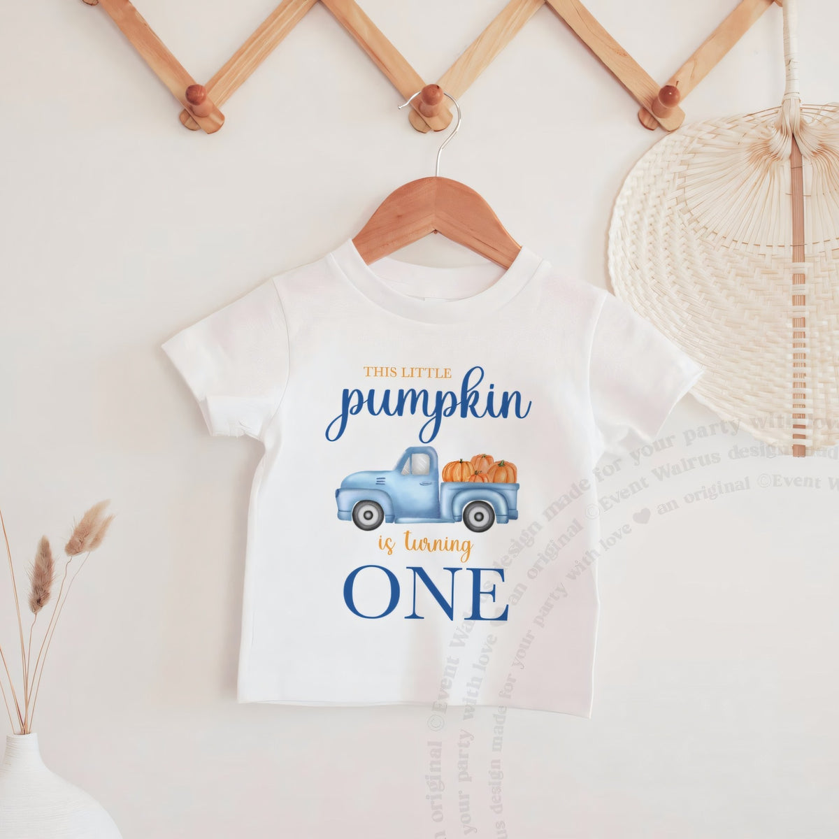 White This Little Pumpkin Is Turning One baby tee hanging on a wooden peg rail.
