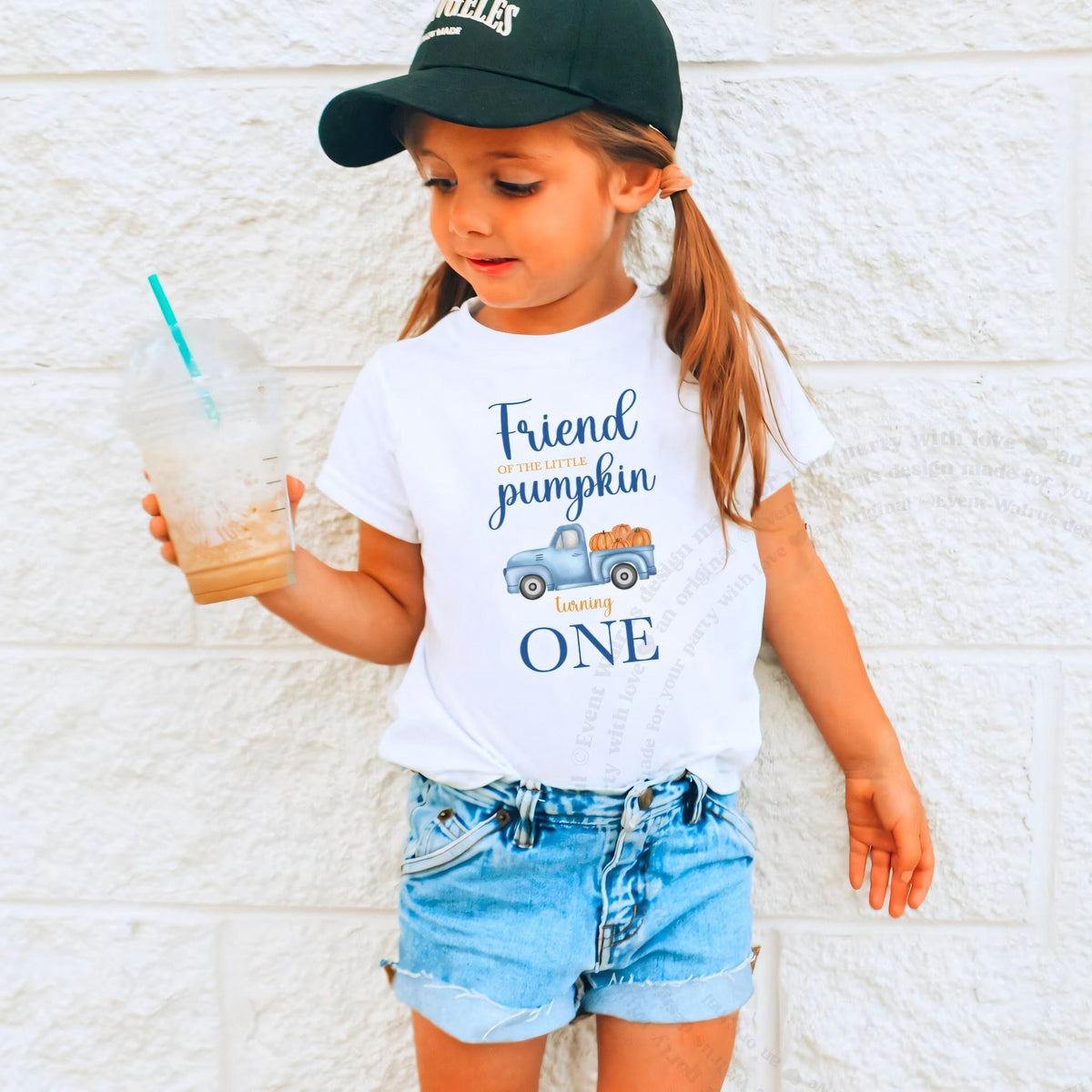 Girl in a ball cap holding a drink in a white Friend of the Little Pumpkin shirt.