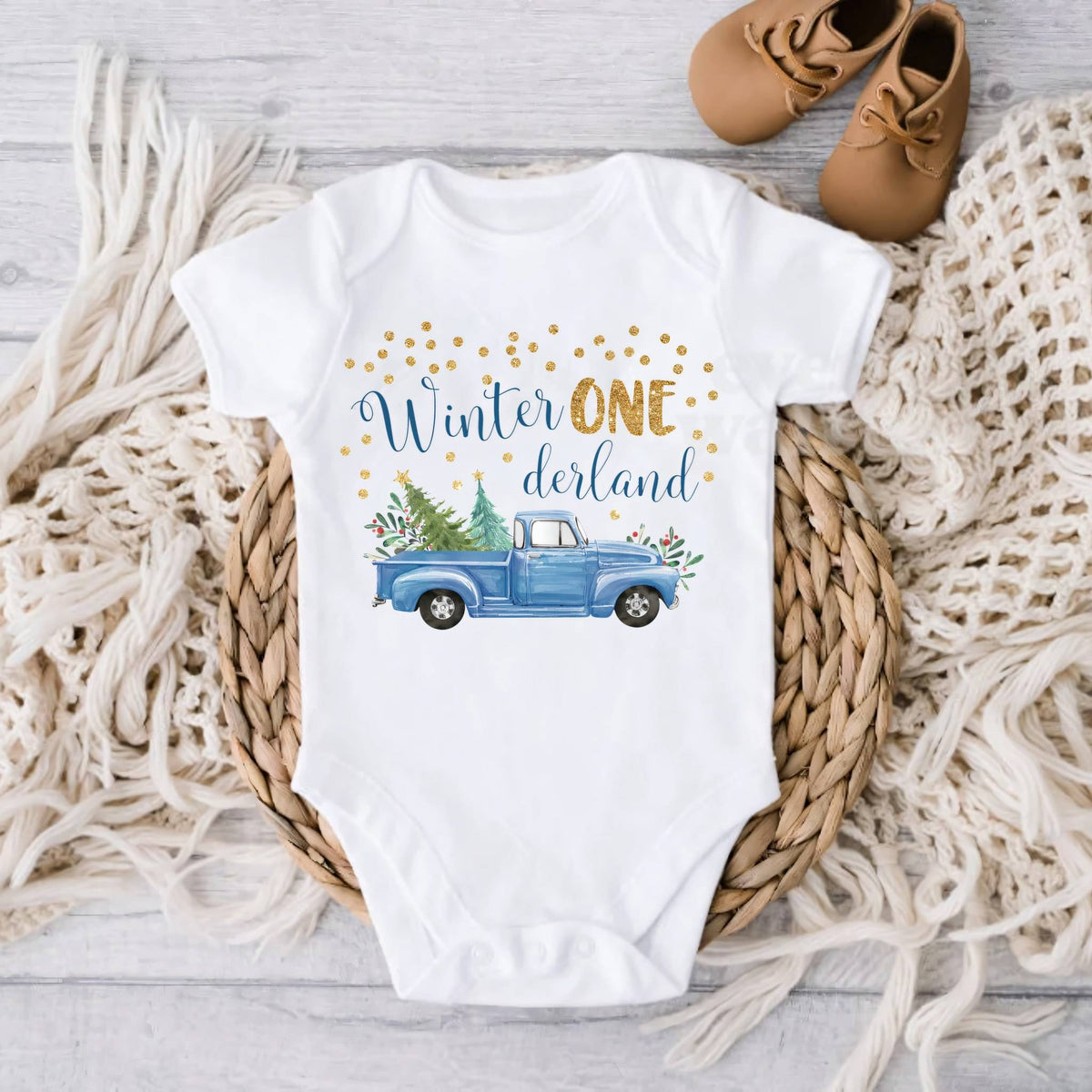 White blue truck Winter ONE derland bodysuit on gray wood with a knit blanket and baby shoes.