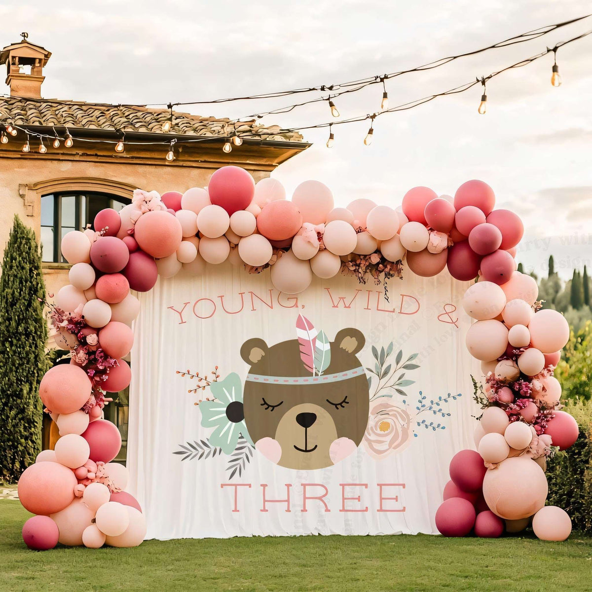 Young wild and three backdrop on a lawn by a villa with a pink and blush balloon arch.