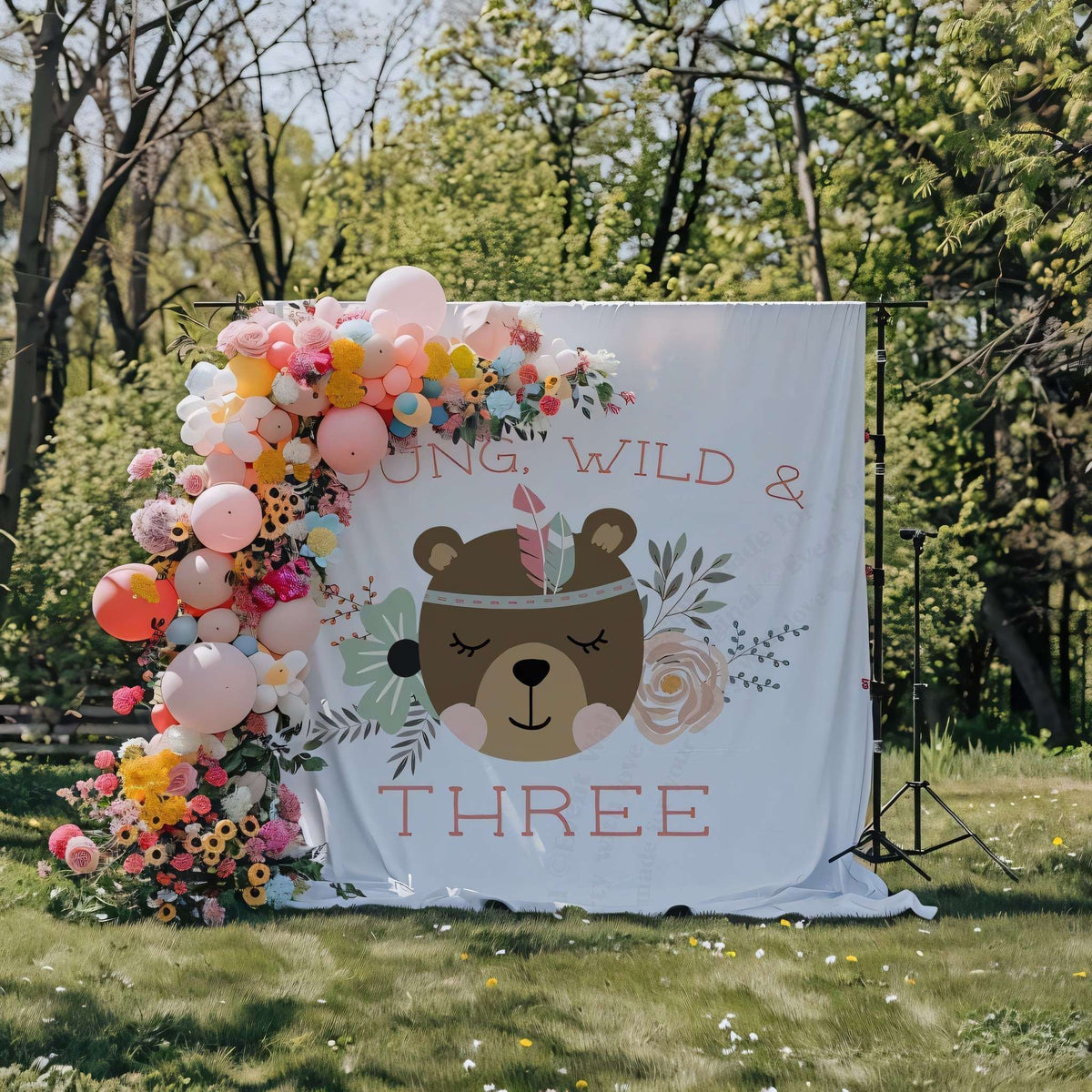Boho bear backdrop in a garden with a wildflower and balloon garland on one side.