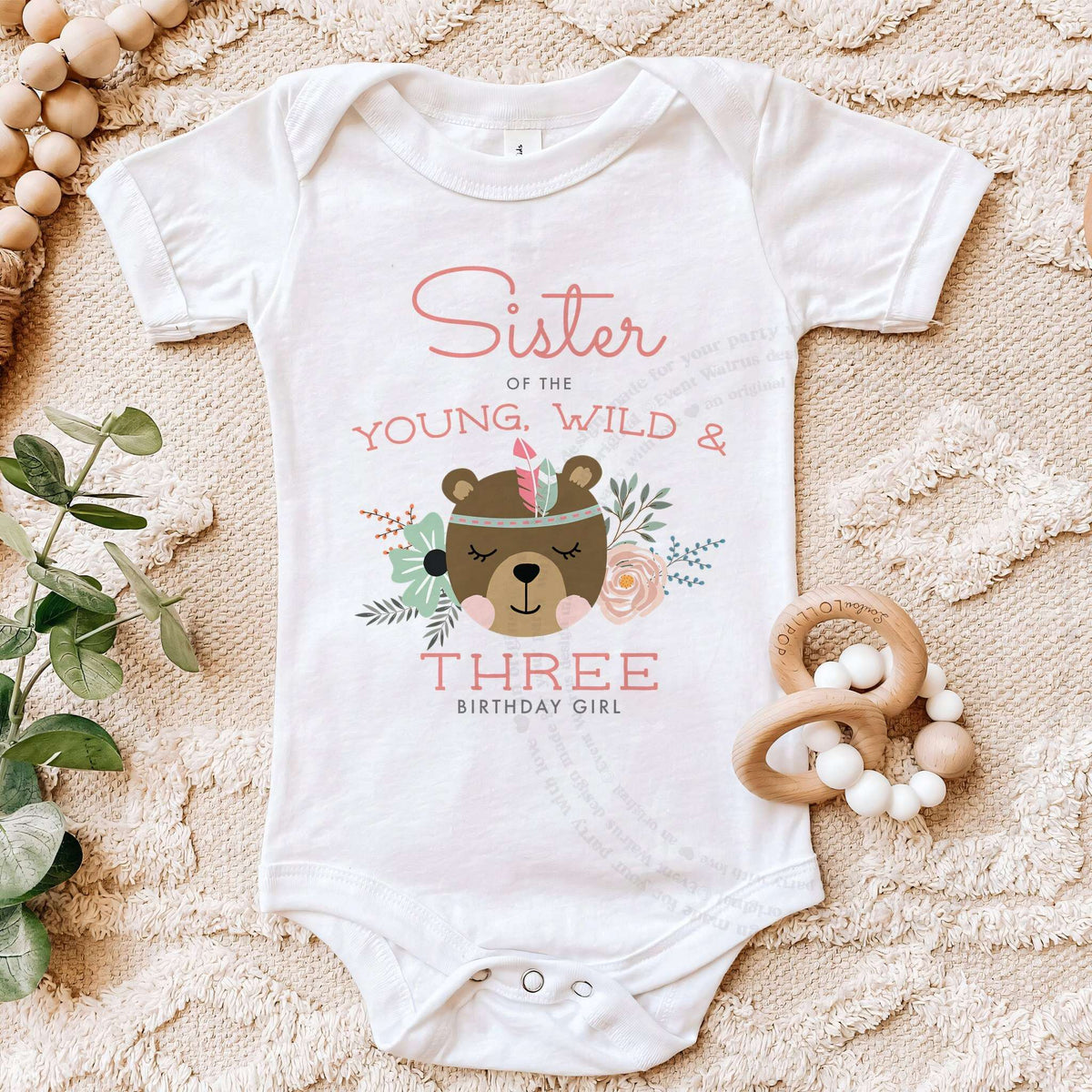 White Sister of the Young, Wild and Three bodysuit on a beige blanket with a wooden teether.