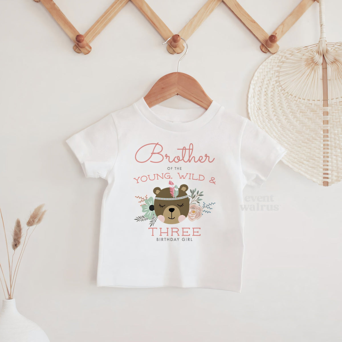 White Brother of the Young, Wild and Three baby tee hanging on a wooden peg rail.