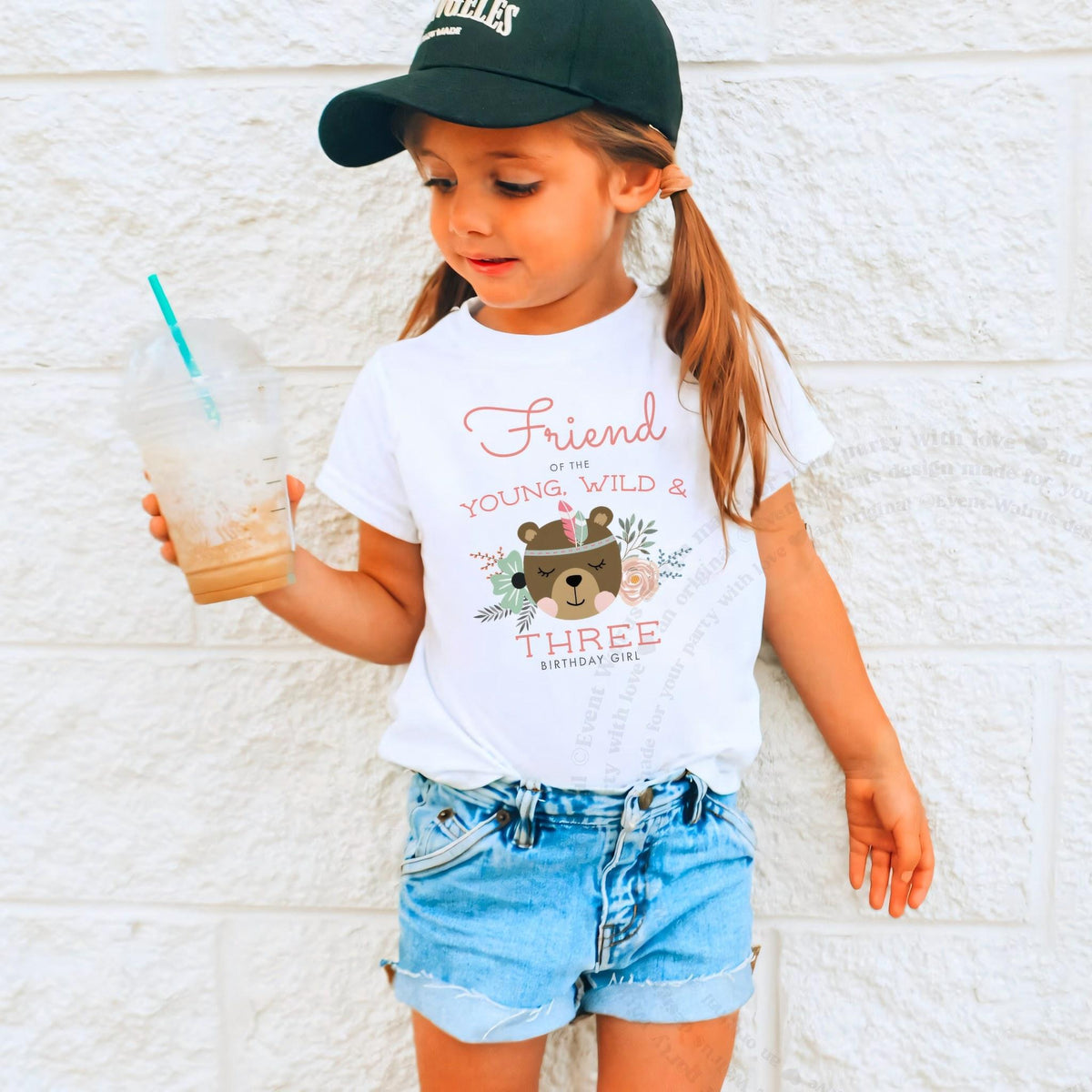 Girl in a ball cap holding a drink in a white Friend of the Young, Wild and Three shirt.
