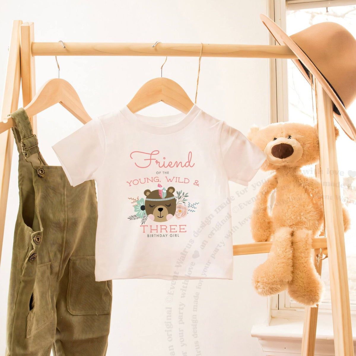 White Friend of the Young, Wild and Three toddler tee on a wooden clothes rack beside a teddy bear.