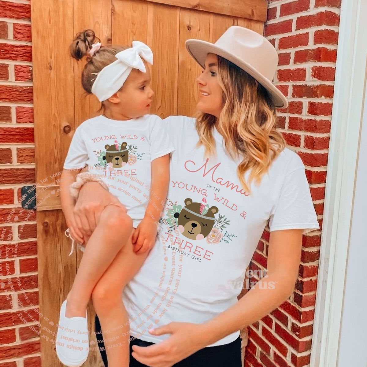 Mom carrying her little girl by a barn door, both in white boho bear Young, Wild and Three shirts.