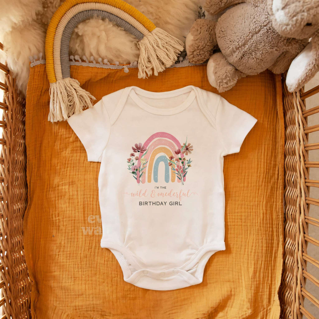 White Wild and Onederful Birthday Girl bodysuit in a wicker basket with a rainbow toy and a teddy bear.