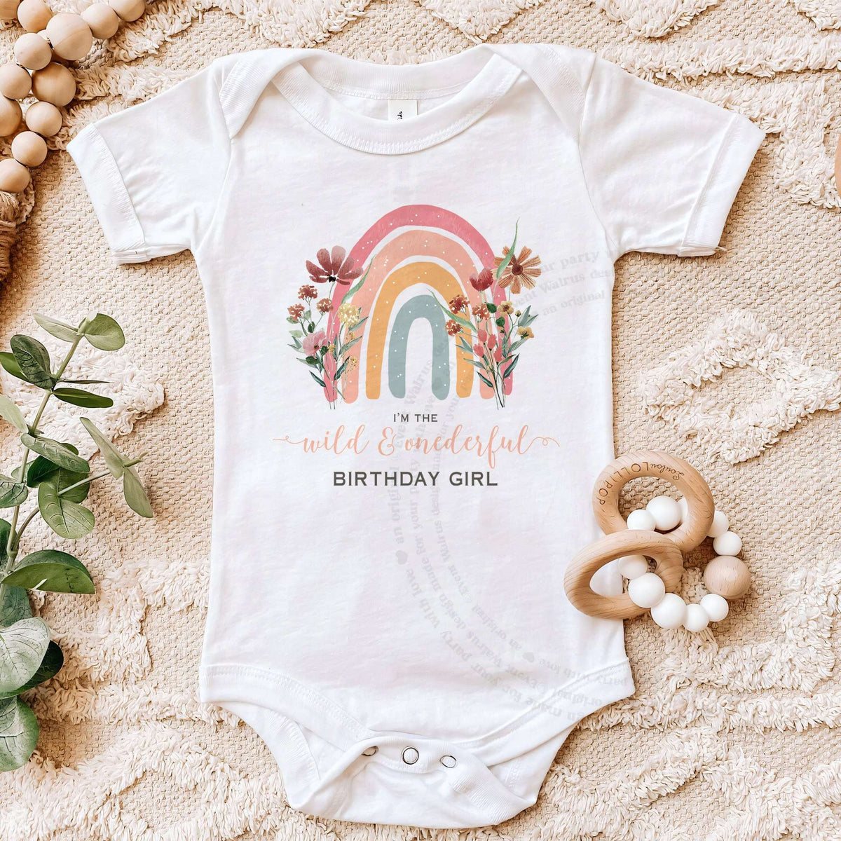 White Wild and Onederful bodysuit on a beige blanket with a wooden teether.