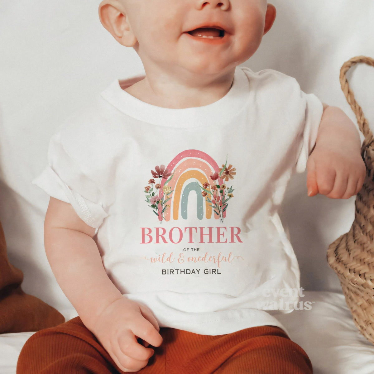 Baby sitting by a woven basket in a white Brother of the Wild and Onederful baby tee.