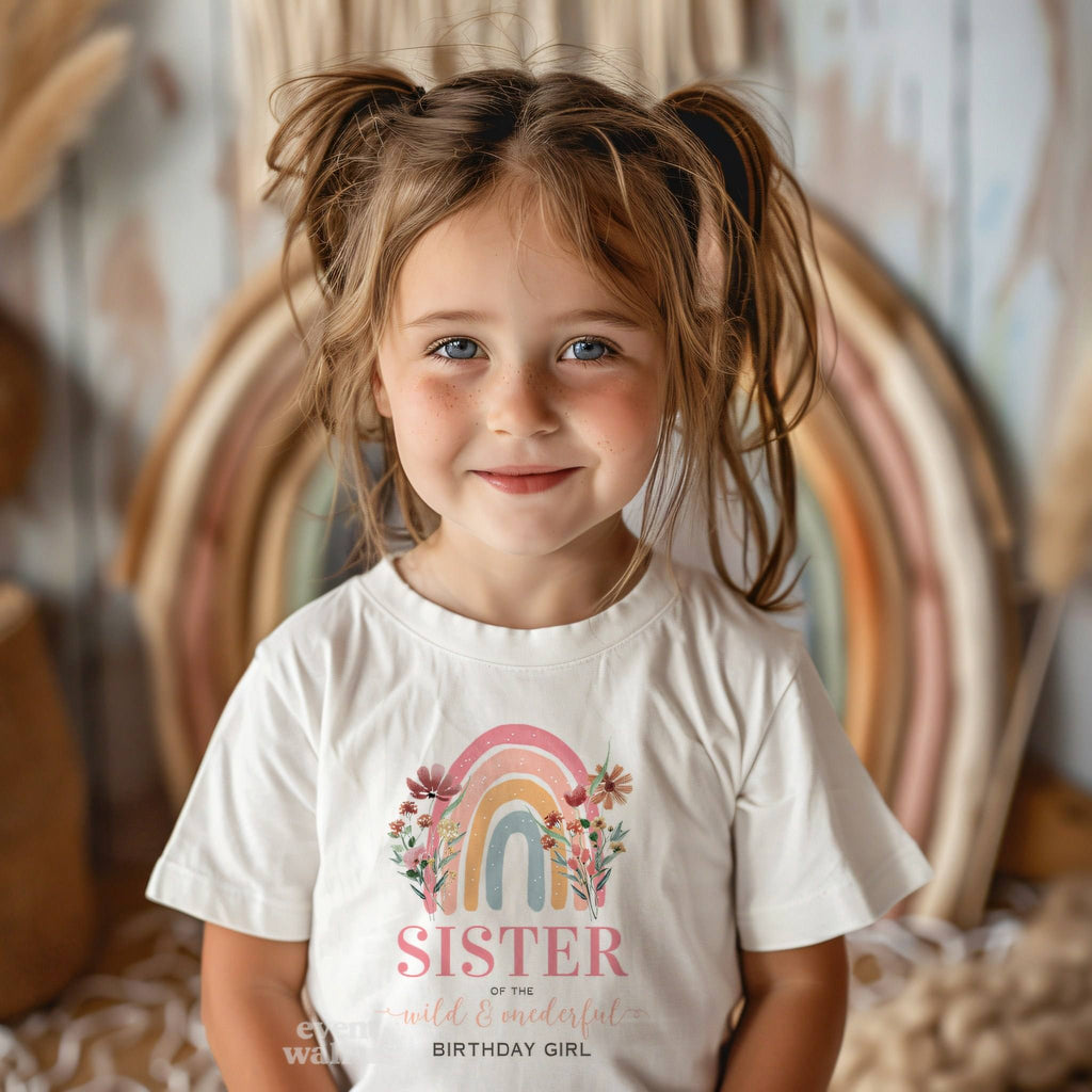 Smiling toddler girl in front of a boho rainbow in a white Sister of the Wild and Onederful tee.