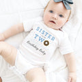 Baby girl with a blue bow lying in a white Sister of the Two Sweet bodysuit.